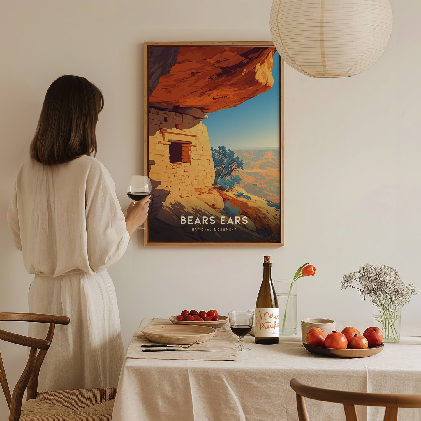 A woman in a white dress holds a glass of wine, admiring a Bears Ears National Monument poster on the wall, with a table set with fruit and wine below.