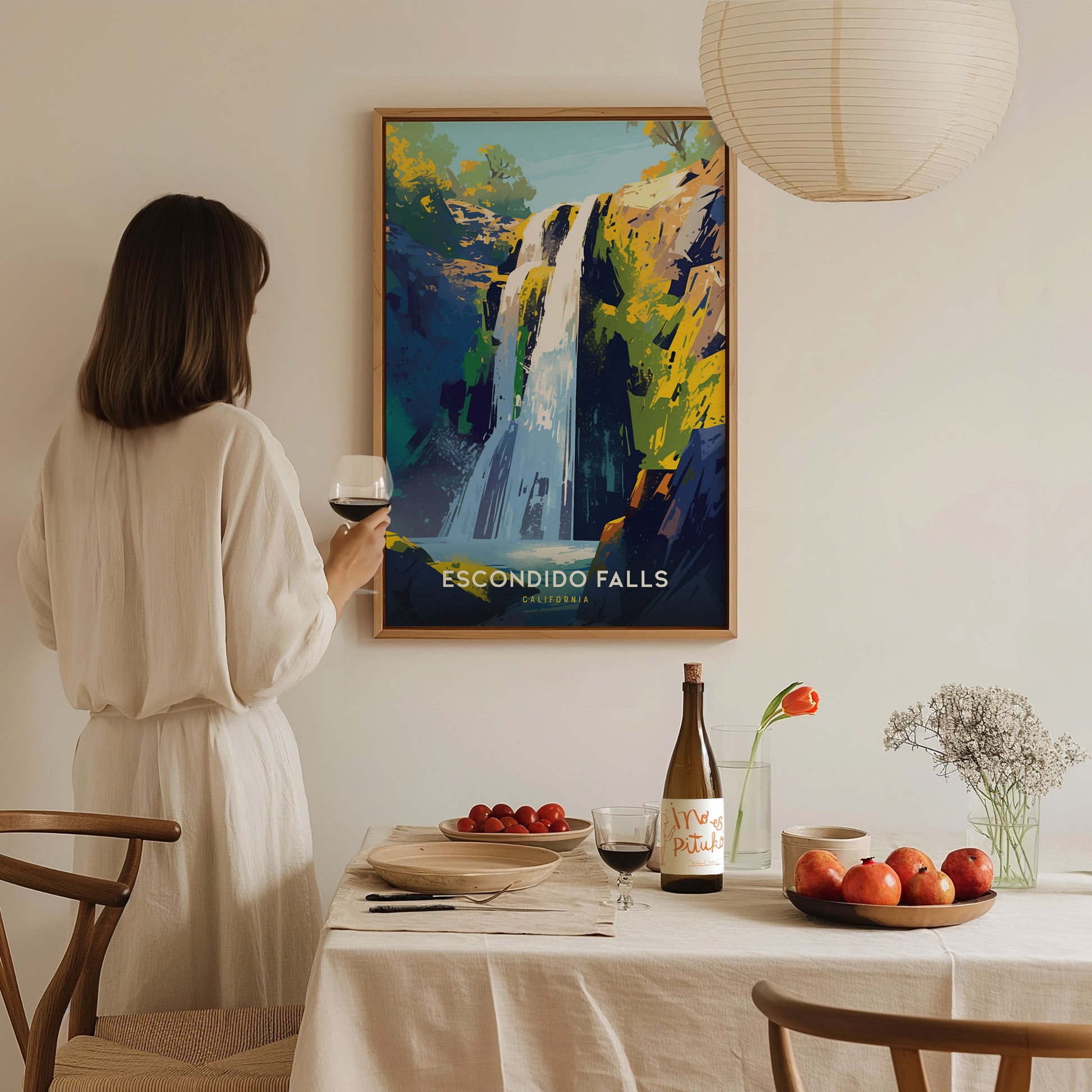 A woman in a white dress holds a glass of wine, admiring a colorful Escondido Falls poster on the wall. A dining table is set with fruit and wine.