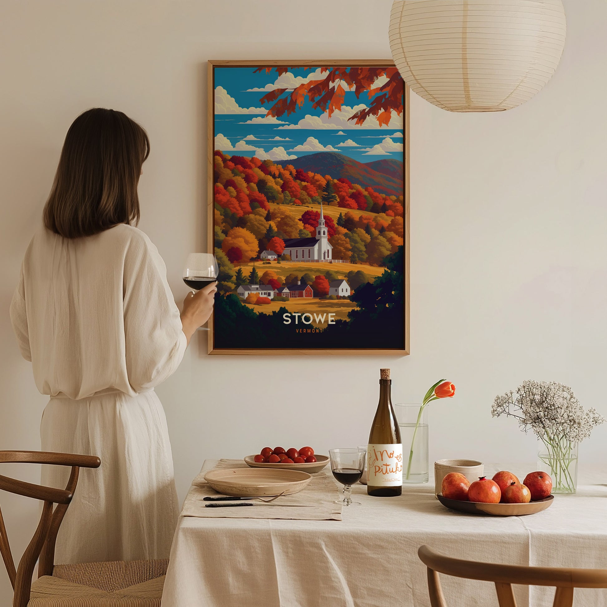 A woman in a white dress holds a wine glass, admiring a Stowe Vermont poster depicting autumn foliage and a church.