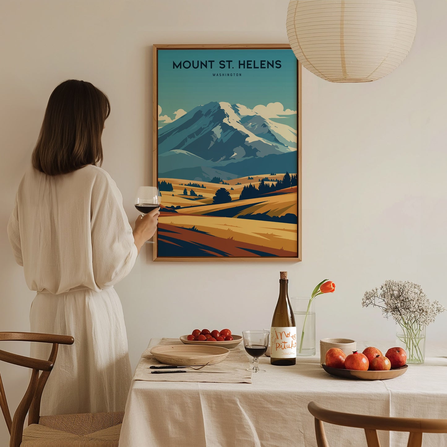 A woman in a white dress admires a Mount St. Helens poster in a dining room with a set table, wine, and fruit.
