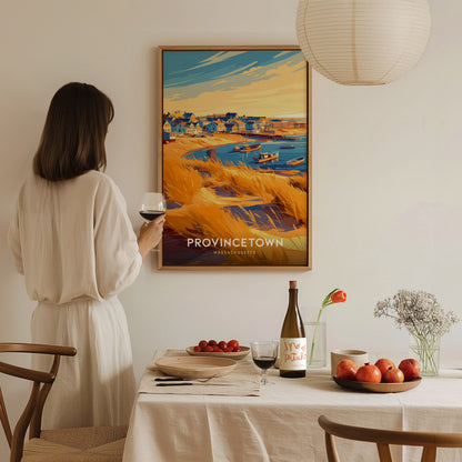 A person in a beige outfit stands beside a dining table, holding a glass of wine, while admiring a vibrant wall art print depicting a coastal scene of Provincetown.