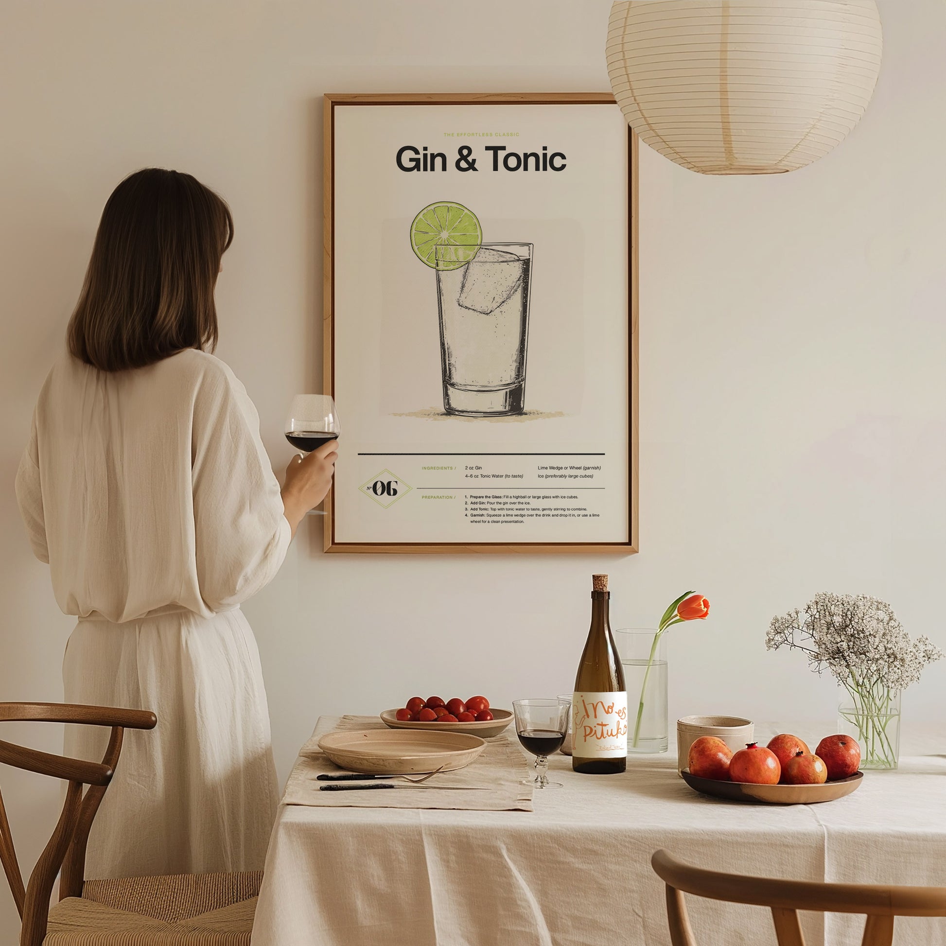 A woman in a white dress holds a wine glass, gazing at a framed Gin & Tonic poster on a wall above a dining table with fruits and flowers.