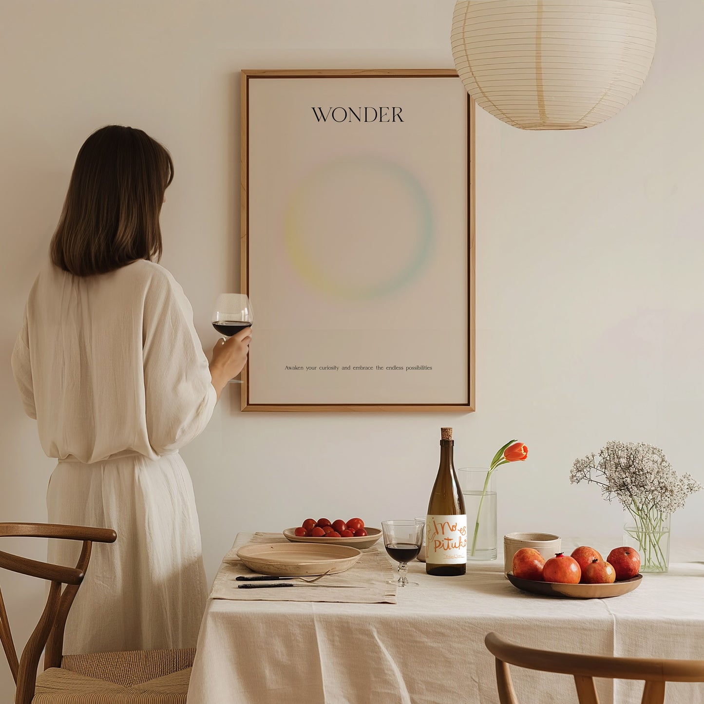 A woman in a light-colored outfit stands holding a glass of wine, gazing at a minimalist art print titled WONDER on the wall. A table set with a bottle, fruit, and flowers complements the calm, inviting atmosphere of the room.