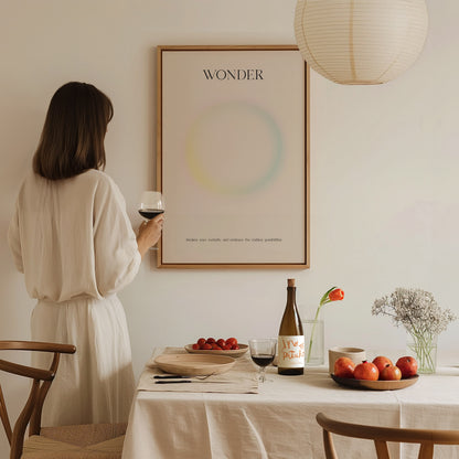 A woman in a light-colored outfit stands holding a glass of wine, gazing at a minimalist art print titled WONDER on the wall. A table set with a bottle, fruit, and flowers complements the calm, inviting atmosphere of the room.