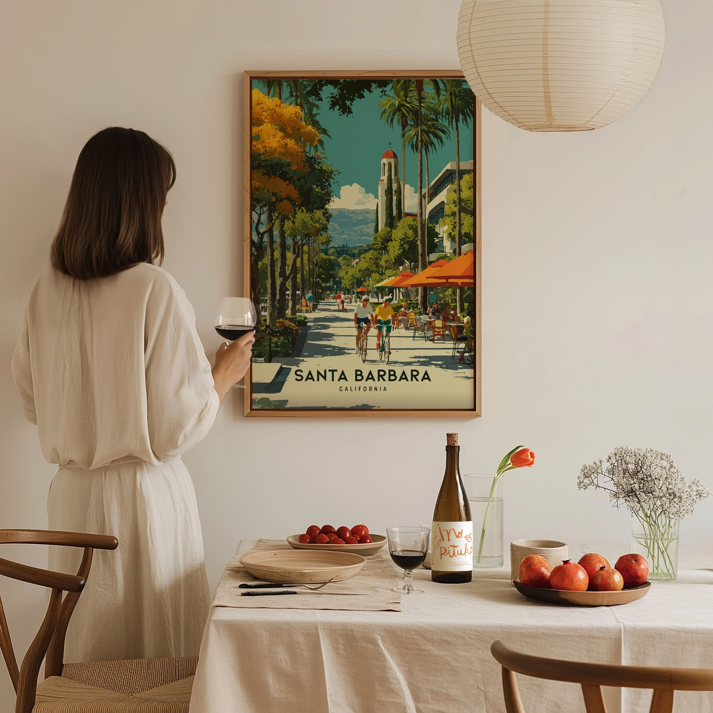 Woman in a white dress standing in a dining room with a framed poster of Santa Barbara on the wall.