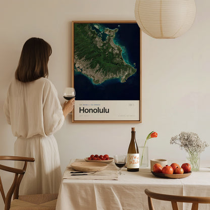 A woman in a white dress stands by a dining table, holding a glass of wine, with a framed map of Honolulu on the wall behind her. The table is set with a bottle of wine, plates, and fresh fruit, creating a cozy dining atmosphere.