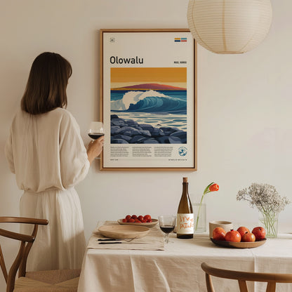 A woman in a white dress holds a glass of wine, admiring a framed Olowalu Maui surf poster on the wall. A table with fruits, wine, and flowers is nearby.