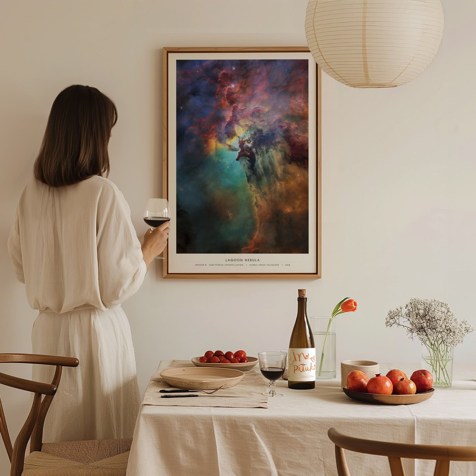 A woman in a white dress stands near a table set with a bottle of wine, glasses, and fruit, gazing at a colorful framed print of the Lagoon Nebula on the wall. The cozy setting features soft lighting and a minimalist decor style.