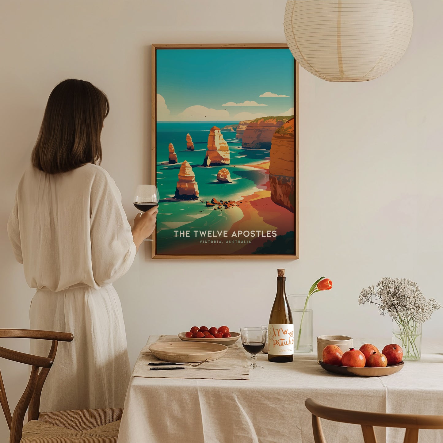 A woman in a white dress holds a glass of wine while admiring a framed poster of the Twelve Apostles. The table is set with fruit, wine, and flowers.