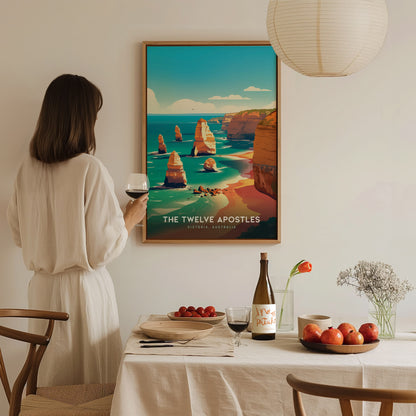 A woman in a white dress holds a glass of wine while admiring a framed poster of the Twelve Apostles. The table is set with fruit, wine, and flowers.