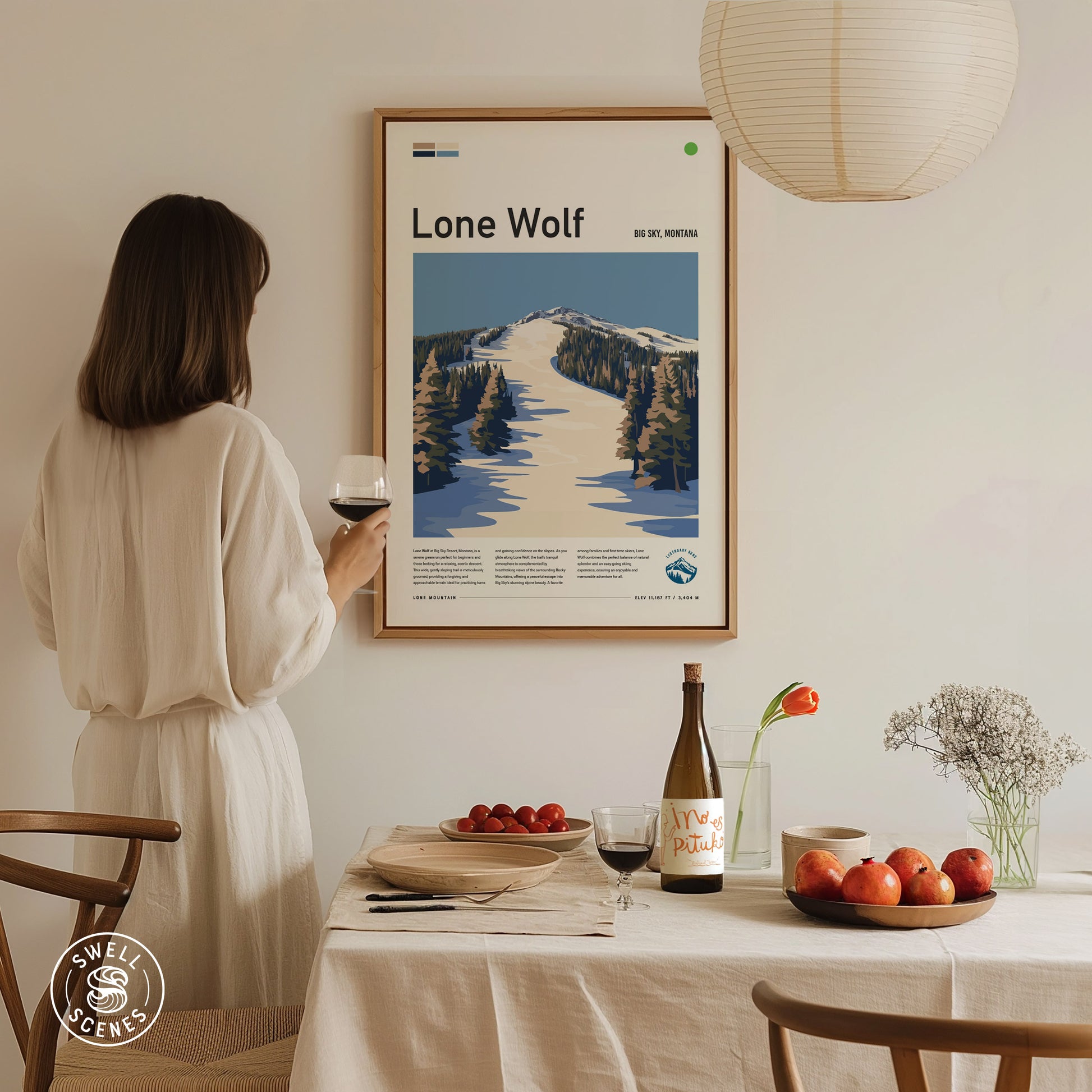 A woman in a white dress holds a glass of wine, admiring a framed Lone Wolf ski poster on the wall. A dining table with fruit and wine is in.