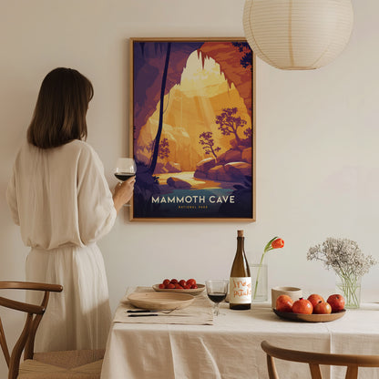 A woman in a white dress holds a wine glass, admiring a framed Mammoth Cave National Park poster. A dining table with fruit, flowers, and wine is in the foreground.