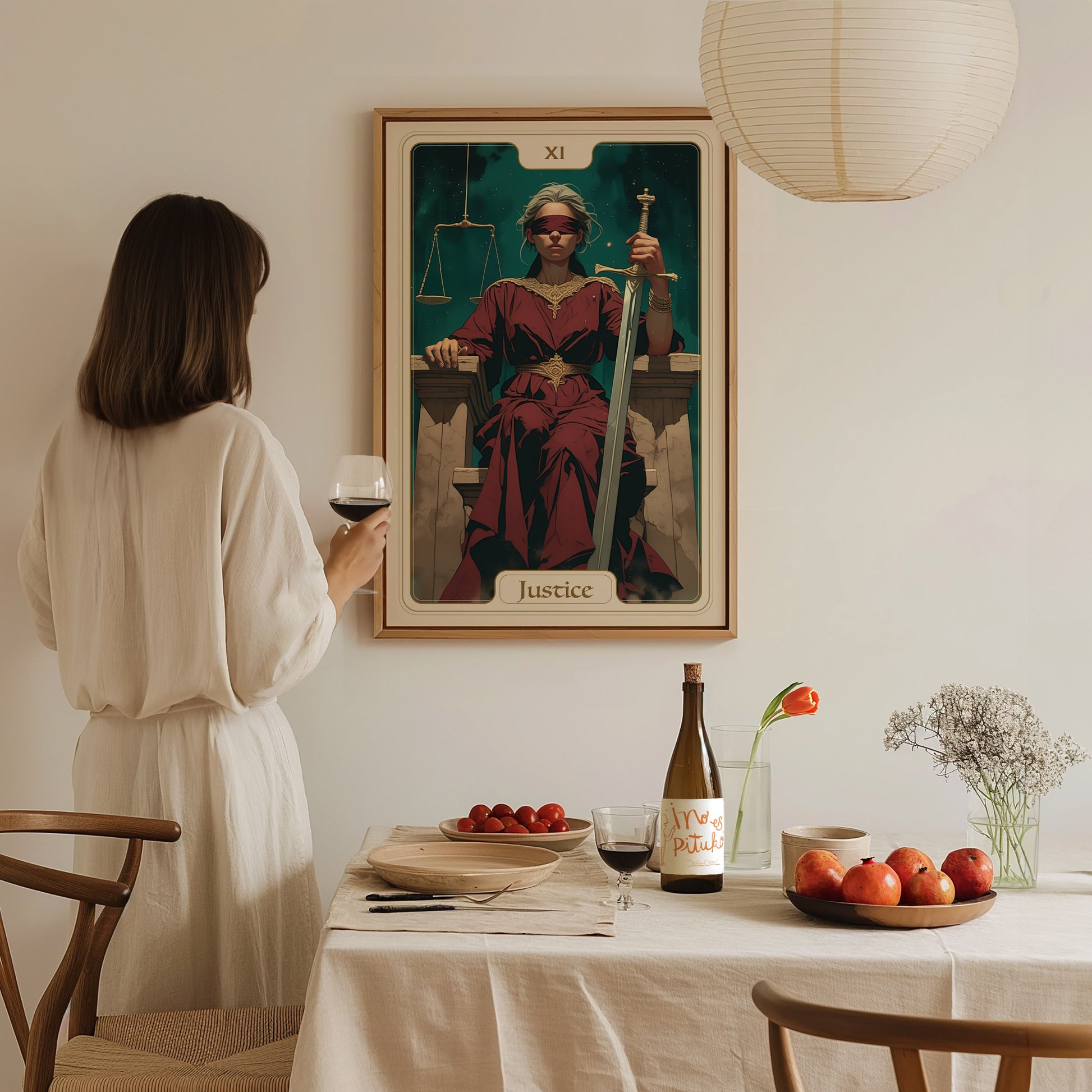 A person in a white dress stands at a dining table, gazing at a framed Justice Tarot Card poster on the wall.