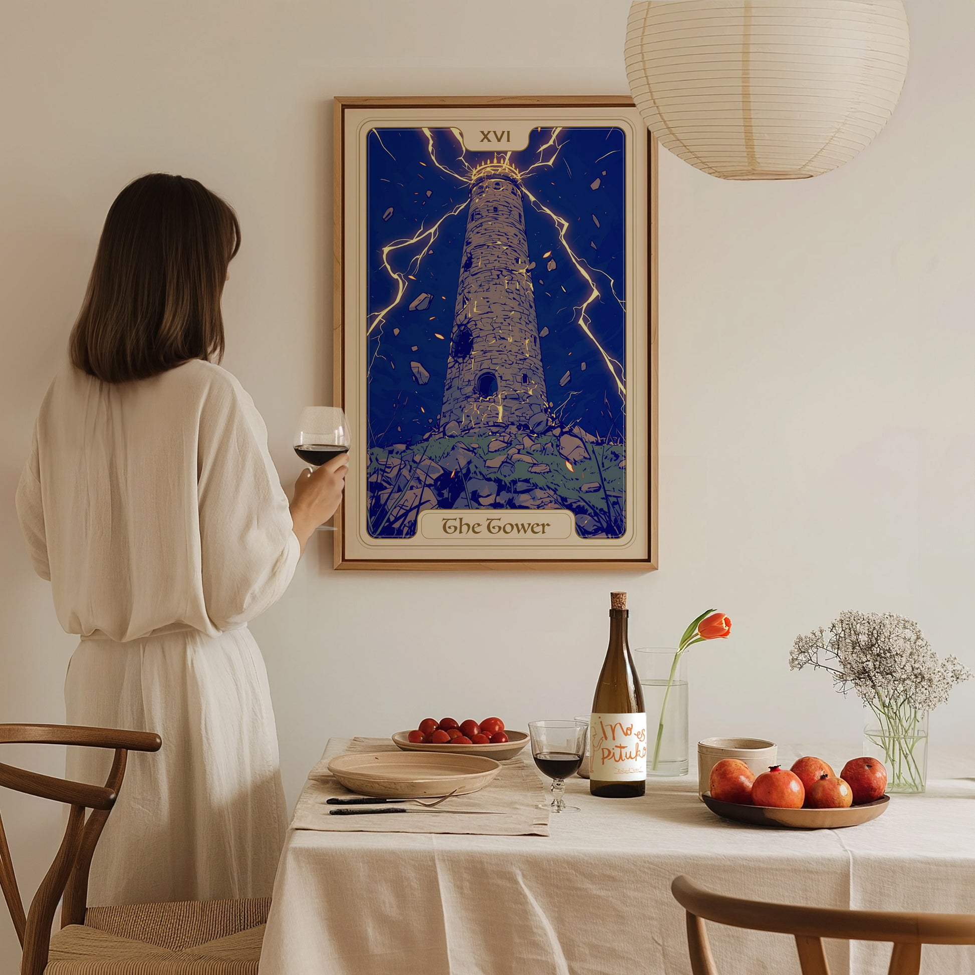 A woman in a white dress stands at a dining table, holding a glass of wine, while gazing at a framed poster of The Tower tarot card on the wall.