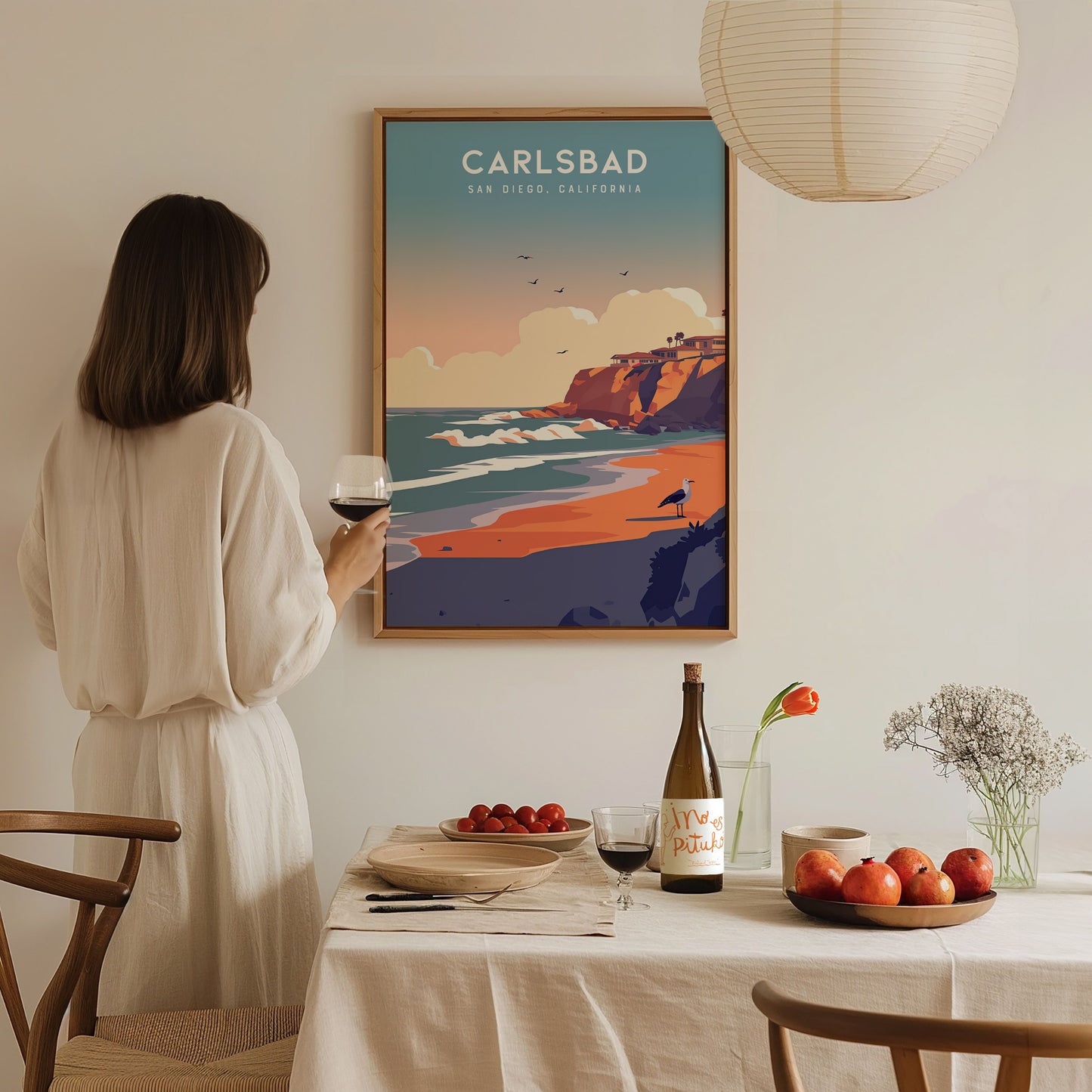 A woman in a white dress holds a glass of wine, admiring a Carlsbad, San Diego poster. A table with fruit, a wine bottle, and flowers is in the foreground.