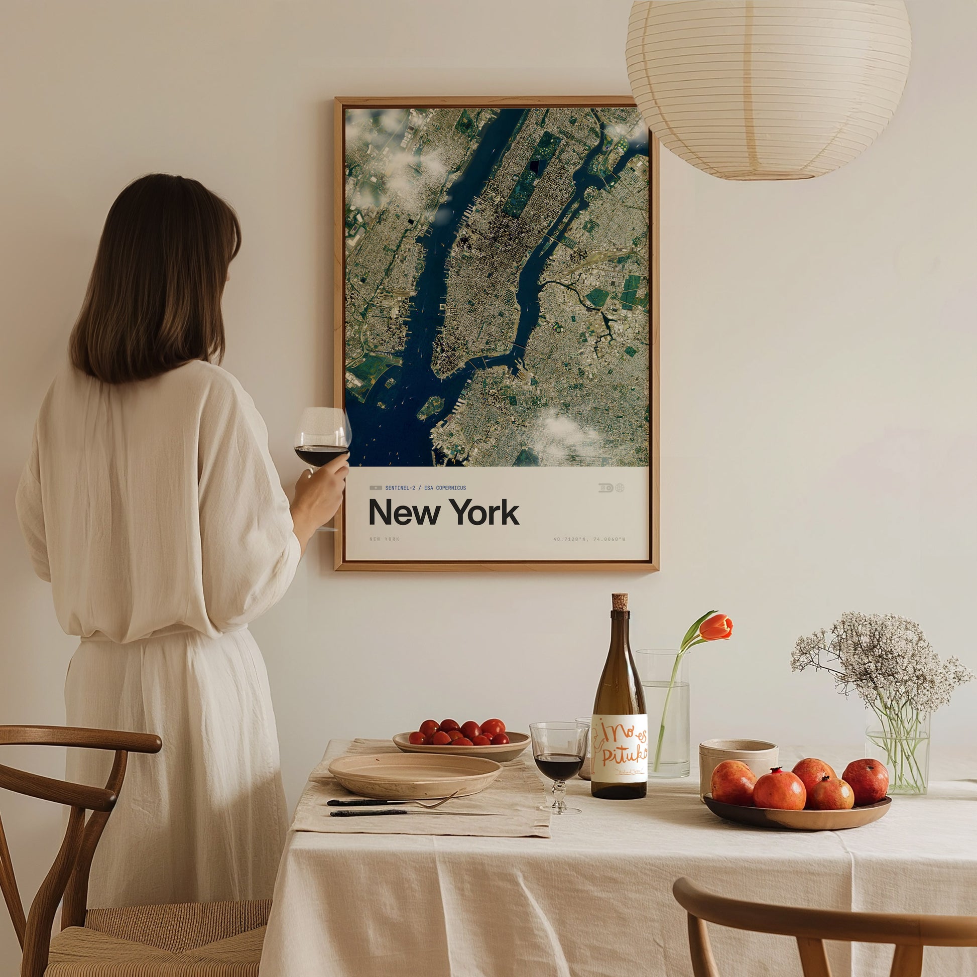 A woman in a white outfit stands holding a glass of wine, gazing at a framed aerial poster of New York City on the wall. A dining table is set with a bottle of wine, a plate of apples, and a vase with flowers, creating a cozy atmosphere.