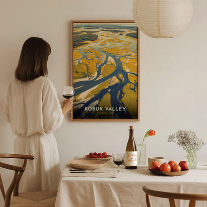 A person in a white dress holds a wine glass, admiring a Kobuk Valley National Park poster. A table with fruits, flowers, and a wine bottle is in the foreground.
