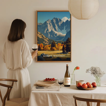 A woman in a white dress holds a glass of wine, admiring a Mount Whitney poster. The table is set with a bottle, red fruits, and flowers.