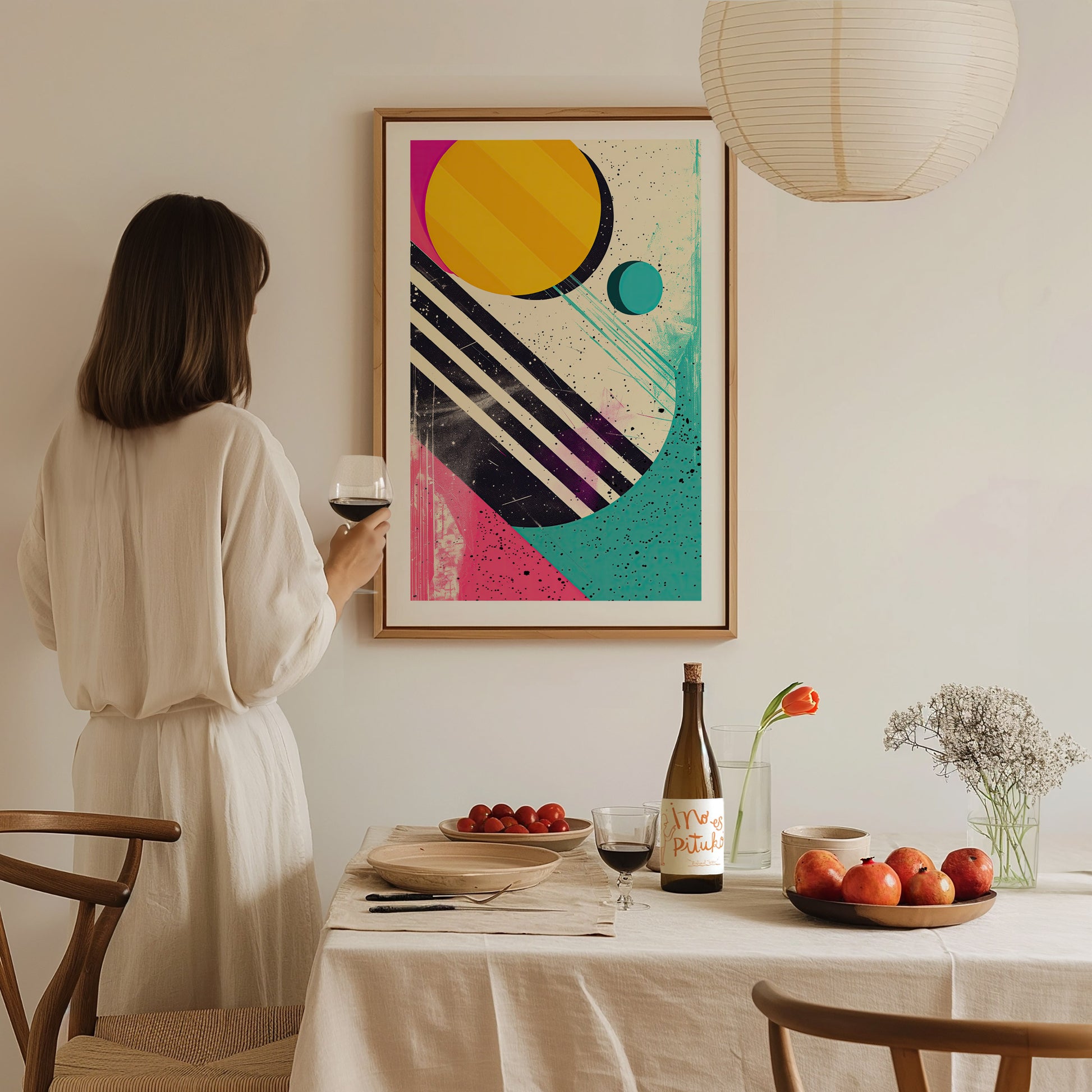 A woman in a white dress holds a glass of wine, admiring a colorful 1980s abstract shapes poster. The table is set with fruit, wine, and flowers.