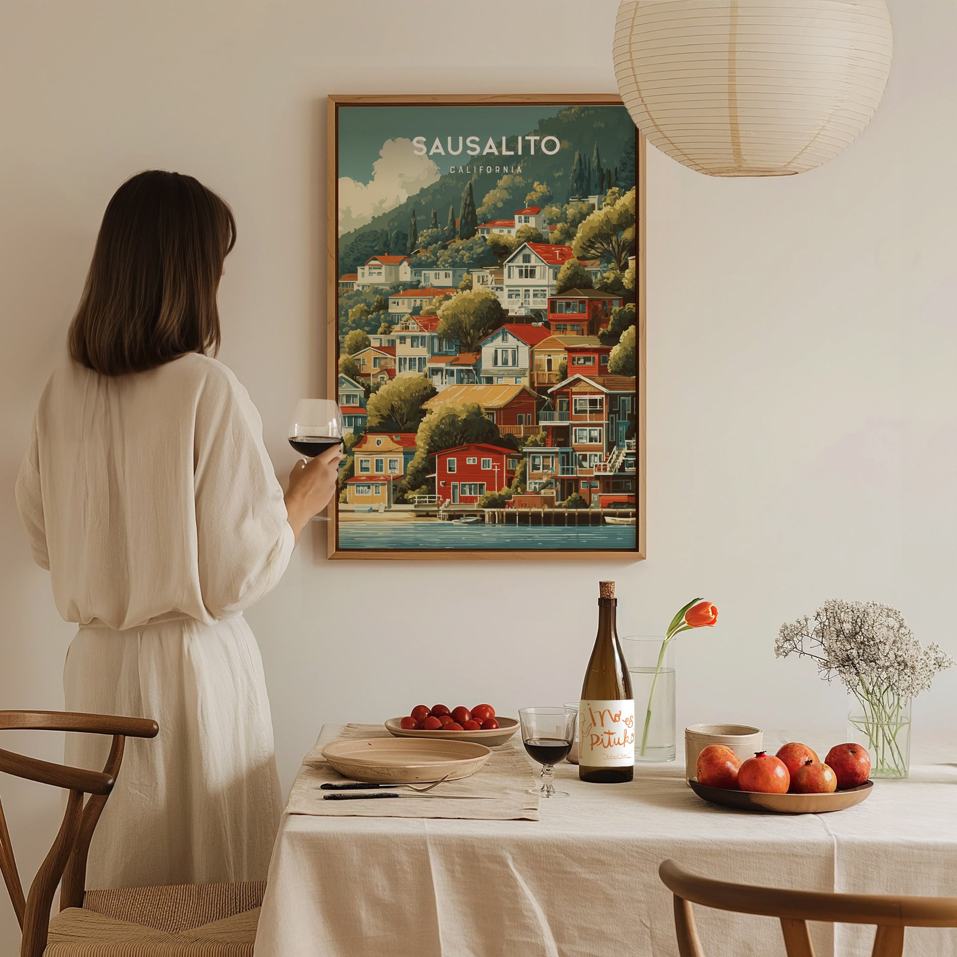 A woman in a white outfit holds a glass of wine while standing next to a dining table adorned with fruits and a bottle.