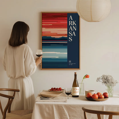 A woman in a white dress holds a glass of wine while looking at a colorful Arkansas poster on the wall. A dining table with fruit and flowers is in.