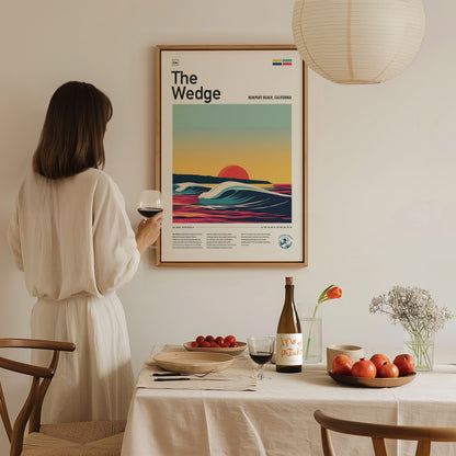 A woman in a white dress holds a glass of wine, admiring a framed surf art poster titled The Wedge on a wall above a table set with fruit and.