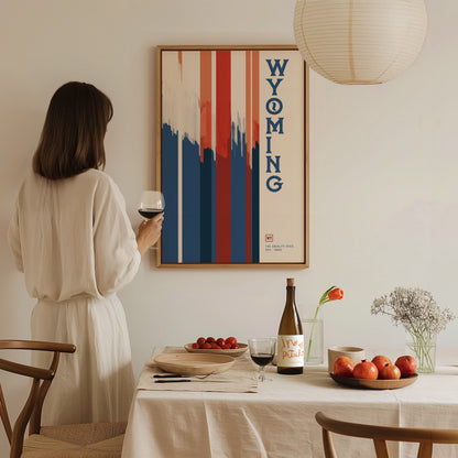A woman in a white dress holds a glass of wine, standing beside a dining table with fruit and a wine bottle.