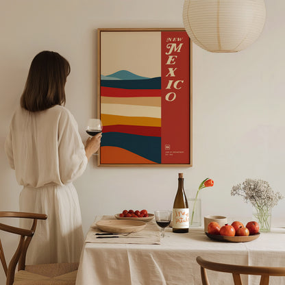 A woman in a white dress admires a colorful New Mexico poster on the wall, holding a glass of wine. A table with fruits and flowers is in the foreground.