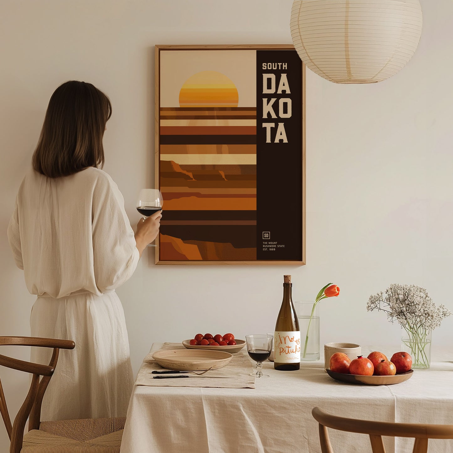 A woman in a white dress holds a wine glass, admiring a South Dakota poster with geometric designs. A dining table with fruit, flowers, and a wine bottle is in.