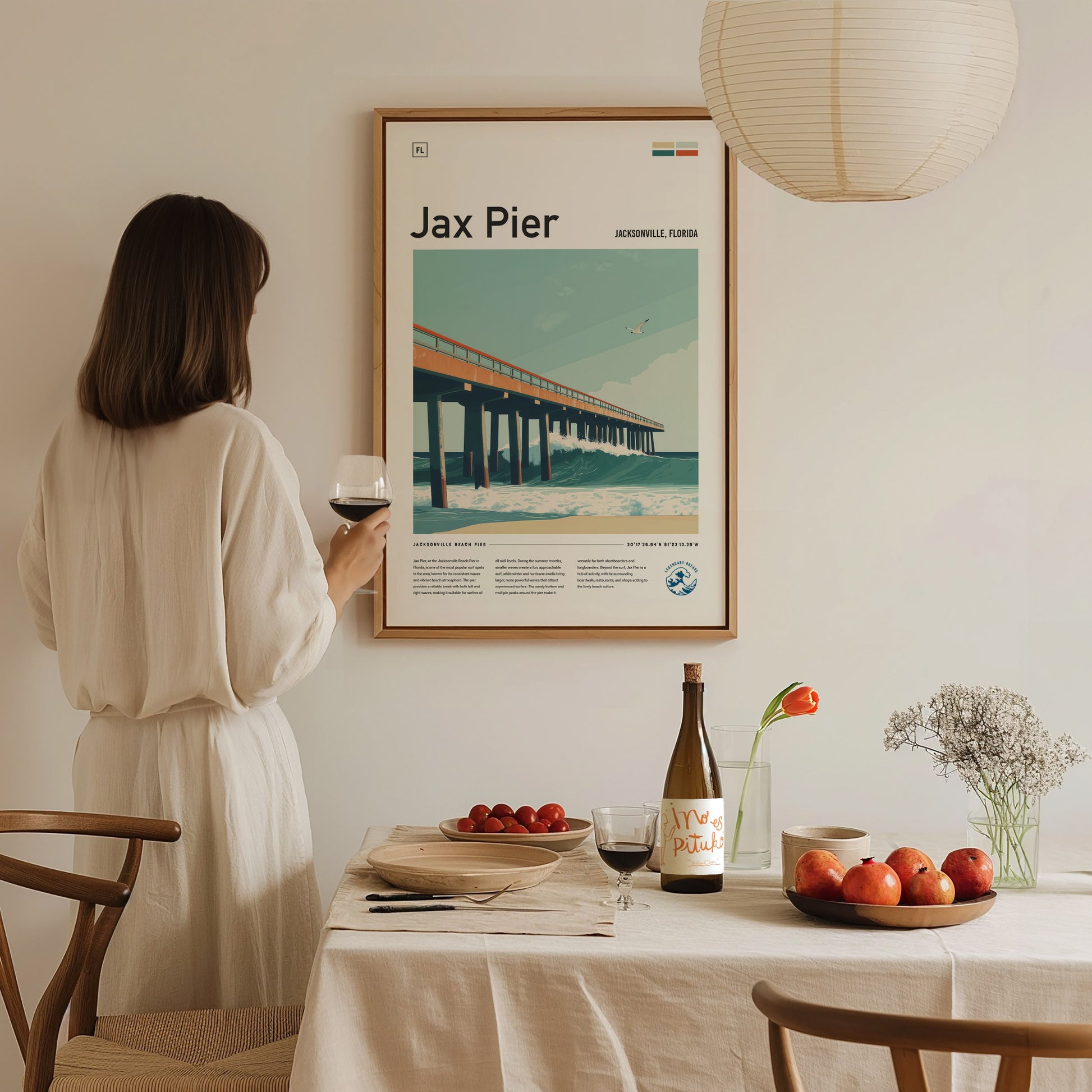 A woman in a white dress admires a framed Jax Pier poster while holding a glass of wine. The table features a bottle, fruits, and flowers, creating an elegant setting.