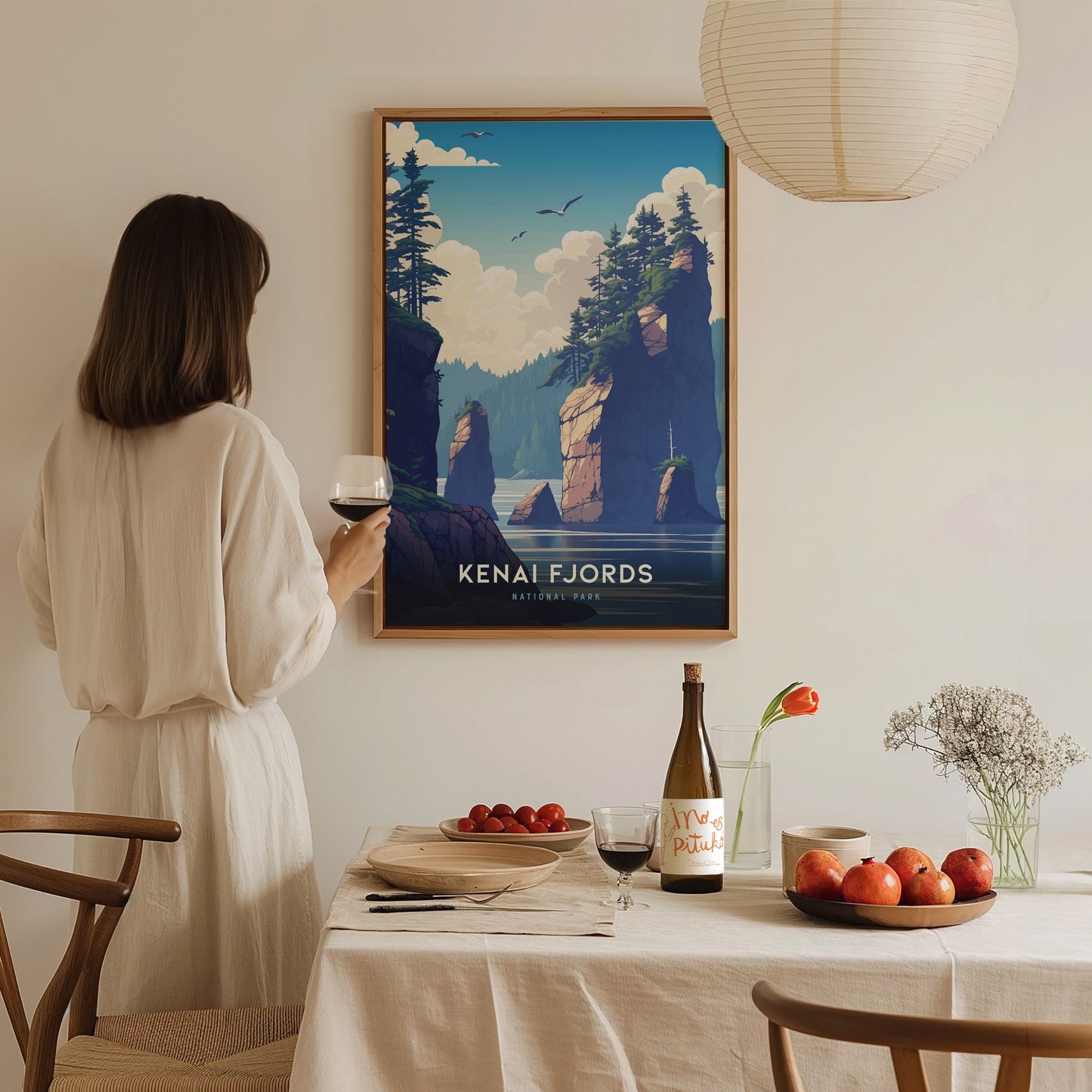 A woman in a white dress holds a wine glass, admiring a Kenai Fjords National Park poster on the wall, while a dining table is set with fruit and flowers.