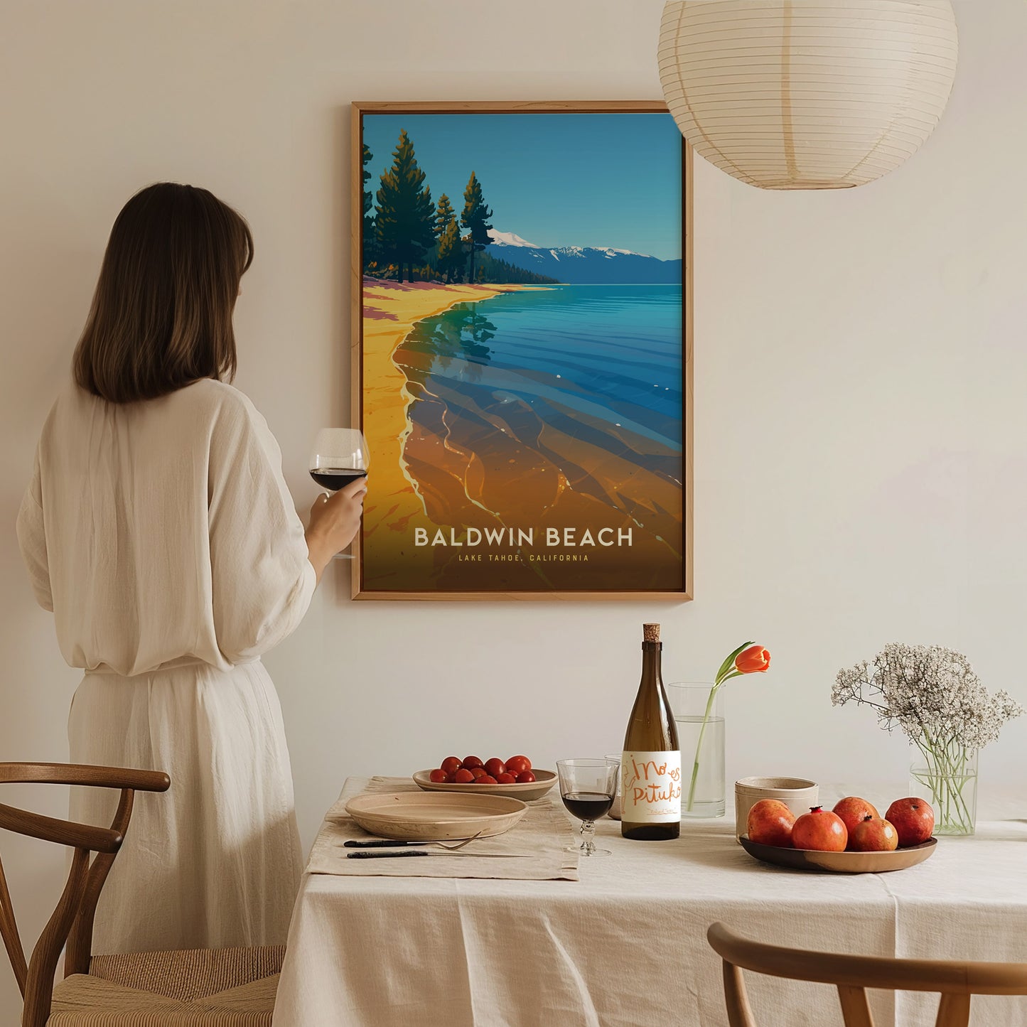 A woman holding a wine glass admires a framed Baldwin Beach, Lake Tahoe poster on the wall. The table is set with a bottle, fruit, and flowers, creating a cozy.