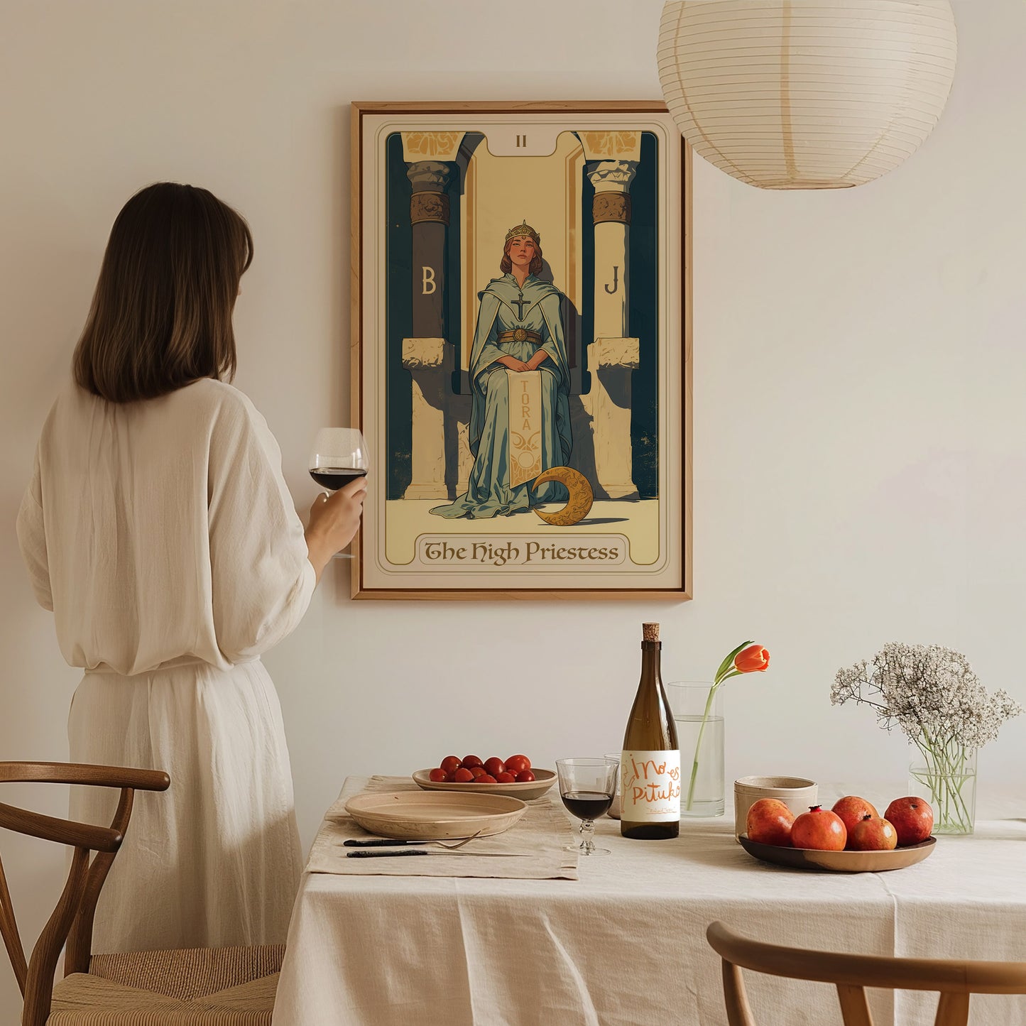 A woman in white holds a glass of wine, admiring a framed High Priestess tarot card poster on the wall. A table with fruit and wine is in the foreground.