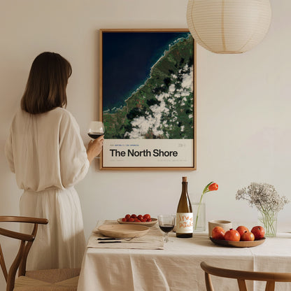 A woman in a white outfit stands holding a glass of wine, gazing at a framed satellite image of Oahus North Shore, which features vibrant greens and deep blues.