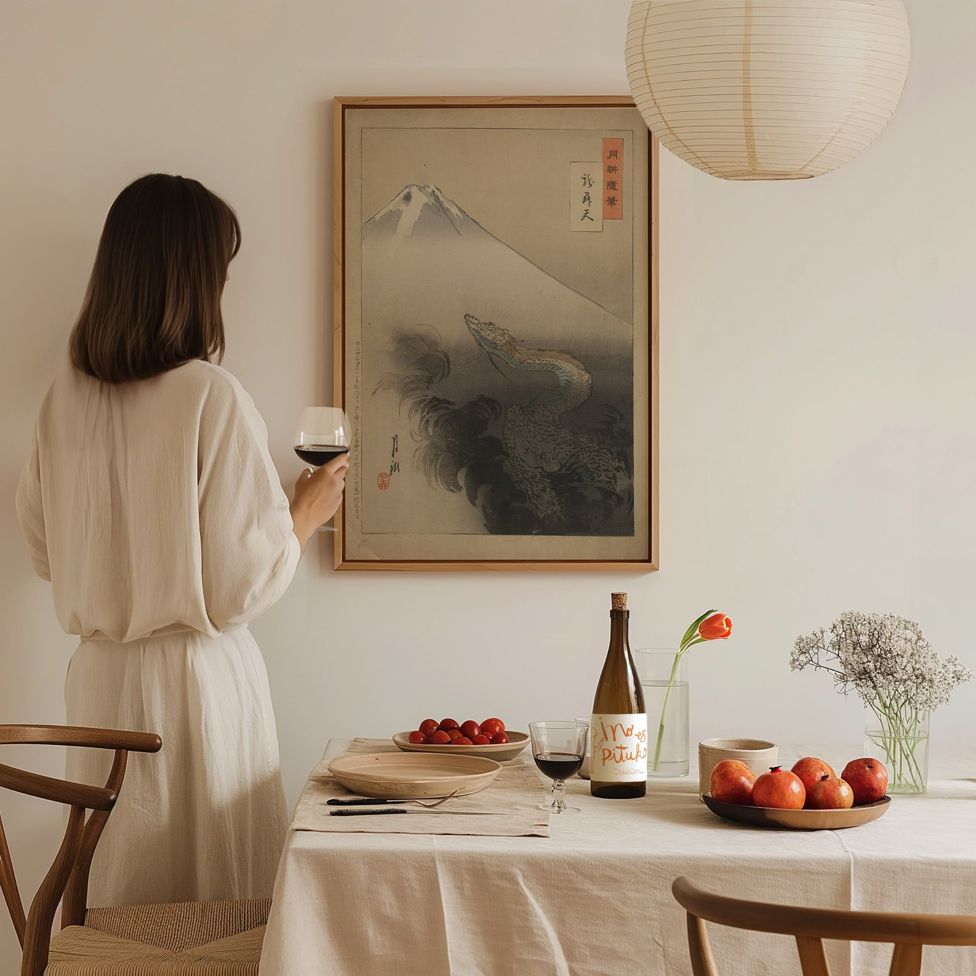 A woman in a white dress holds a glass of wine, admiring a framed dragon poster on the wall. A table with fruits, flowers, and a wine bottle is in.