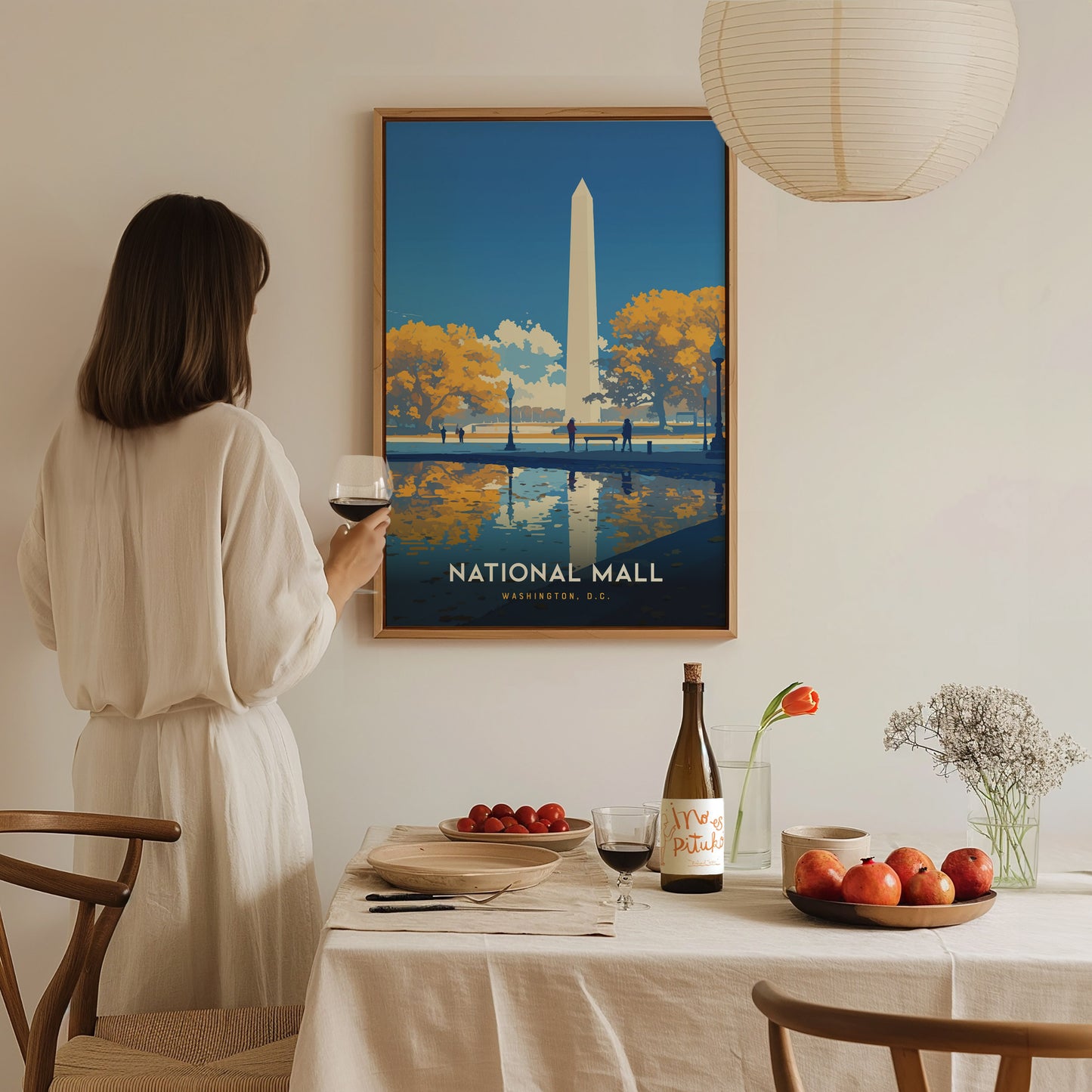 A woman in a white dress holds a glass of wine, admiring a framed National Mall poster. The table is set with fruit, wine, and a vase with flowers.