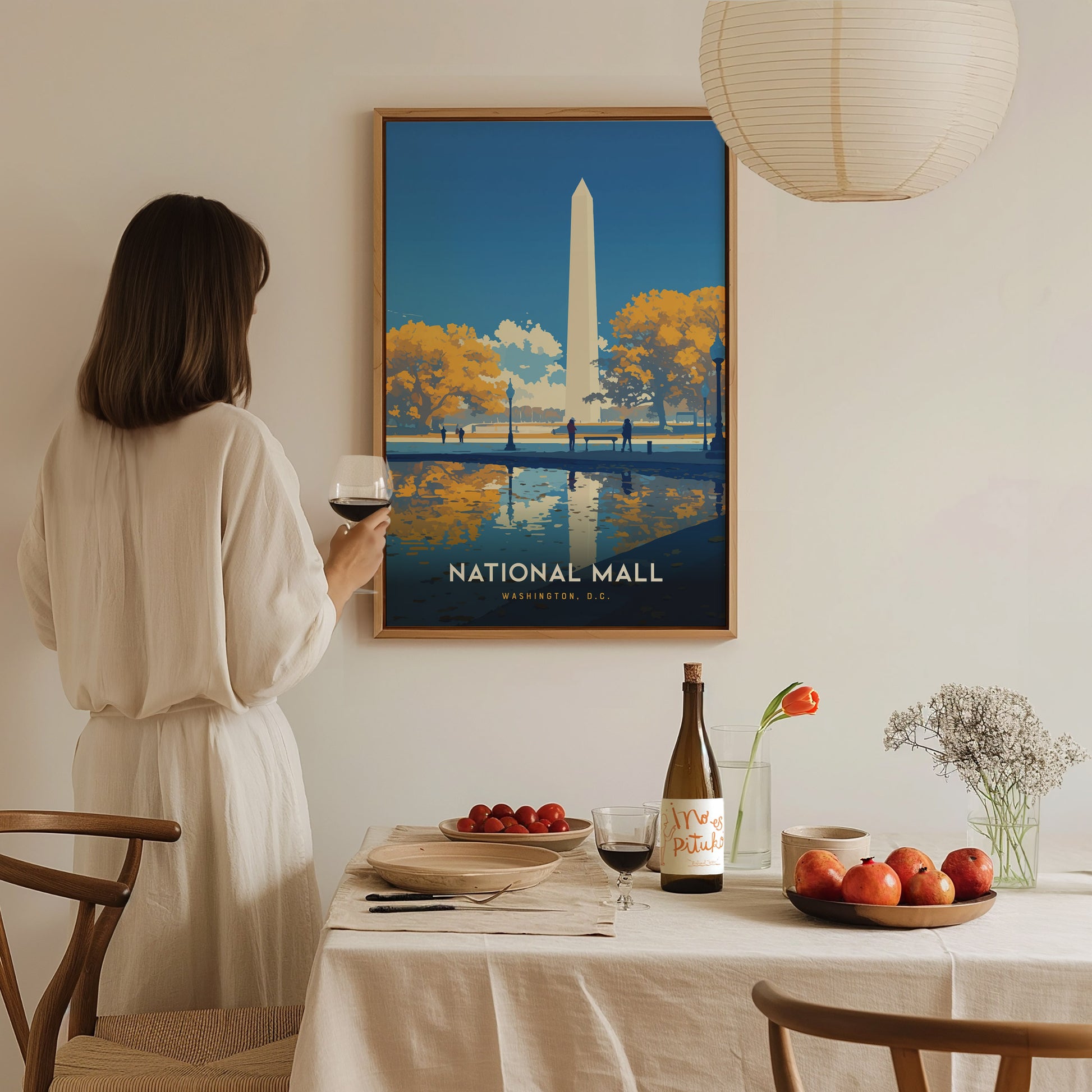 A woman in a white dress holds a glass of wine, admiring a framed National Mall poster. The table is set with fruit, wine, and a vase with flowers.
