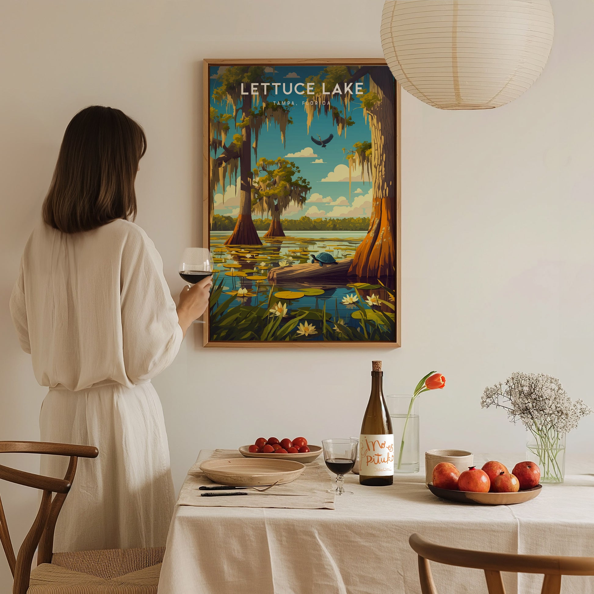 A woman holding a glass of wine admires a framed Lettuce Lake Park poster on a wall above a dining table set with fruit, flowers, and a wine bottle.