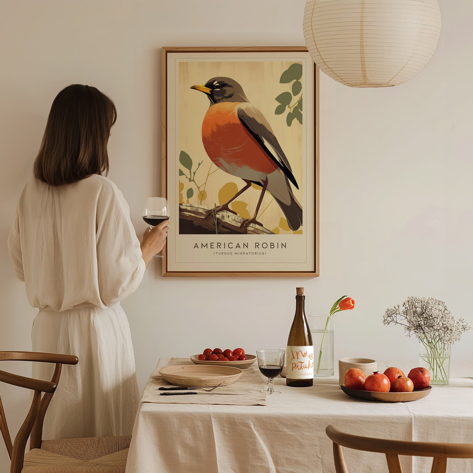 A woman in a white dress admires an American Robin art print on the wall. A dining table below features wine, fruit, and a vase with flowers.