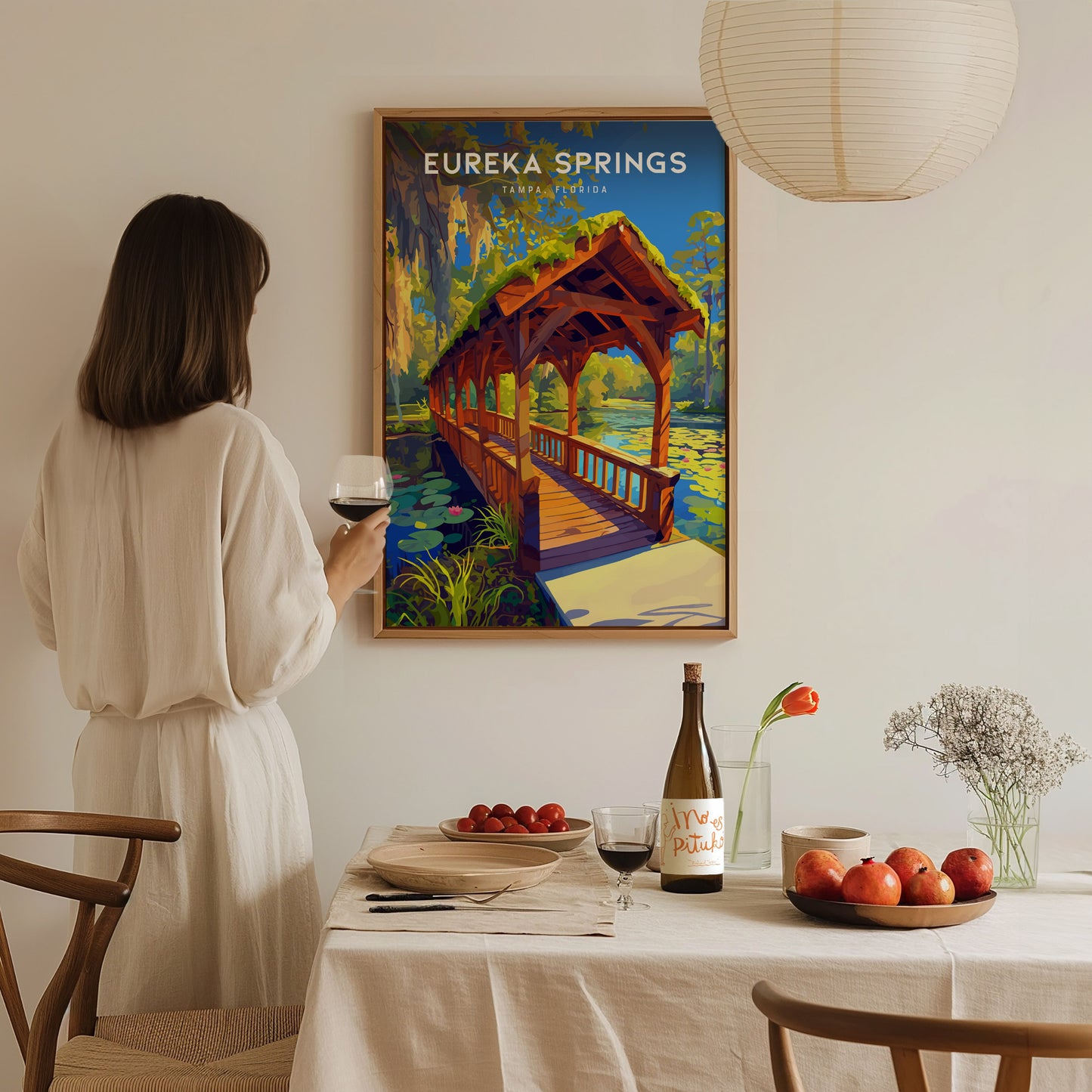 A woman in a white dress holds a wine glass, admiring a framed Eureka Springs poster featuring a wooden bridge.