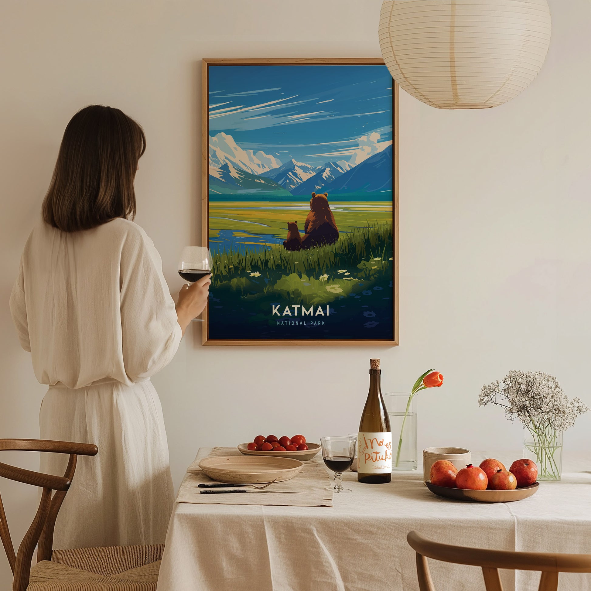 A woman in a white dress admires a Katmai National Park poster featuring bears and mountains. A dining table is set with wine, fruits, and flowers in the foreground.