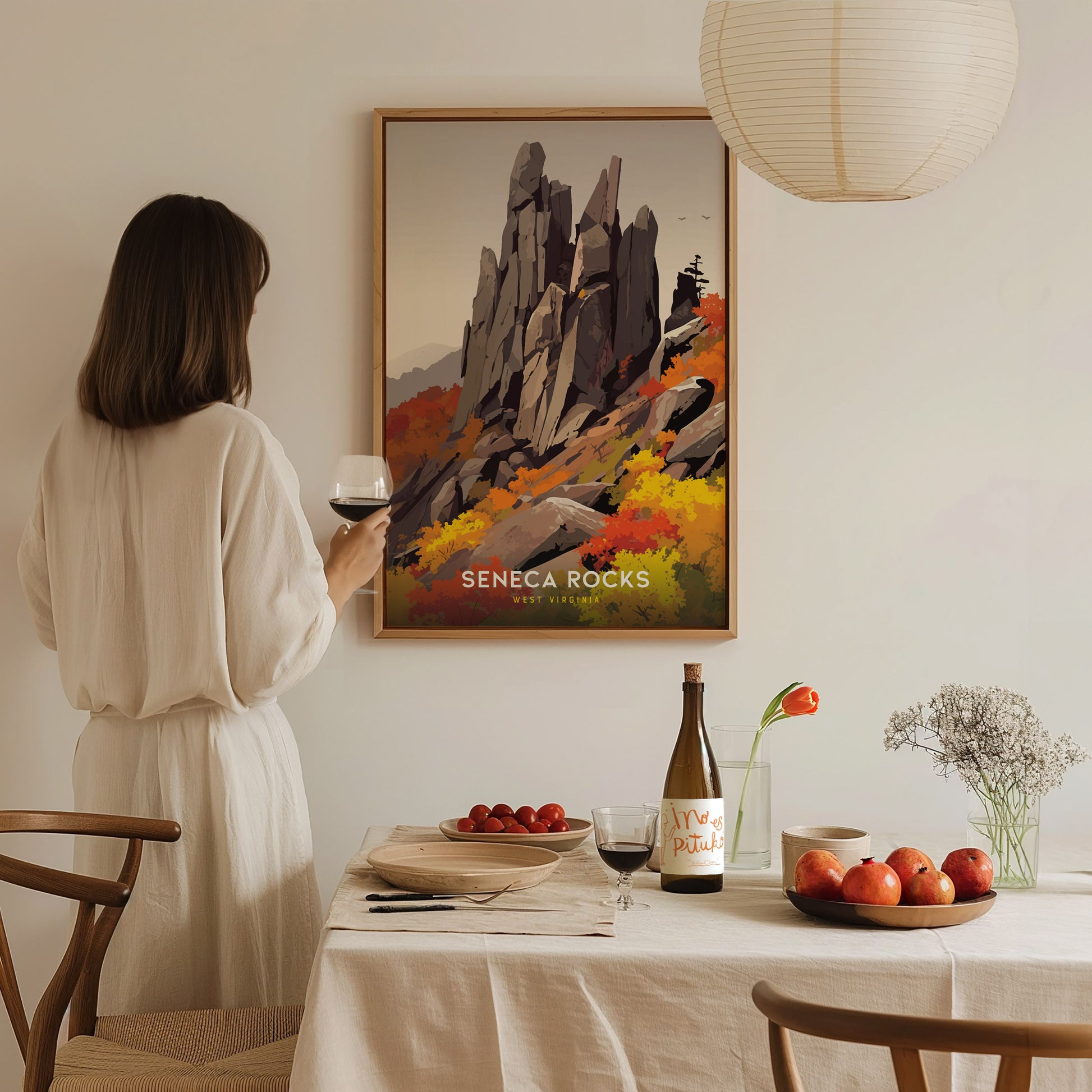 A woman in a white dress holds a glass of wine while admiring a Seneca Rocks poster. The table is set with fruit, wine, and flowers, creating a cozy atmosphere.