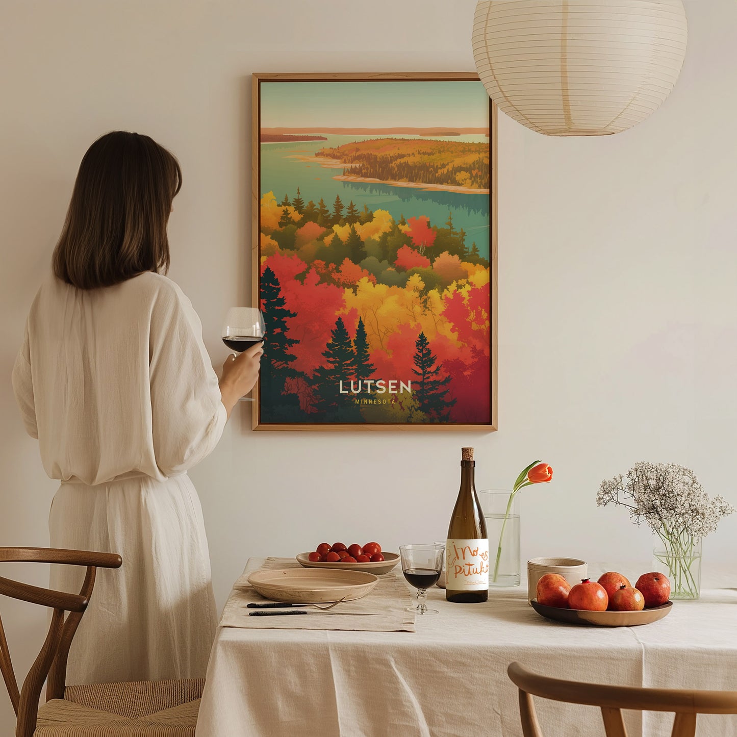 A woman in a white dress holds a wine glass, admiring a colorful Lutsen Minnesota poster on the wall. A table with fruit and a bottle is in the foreground.