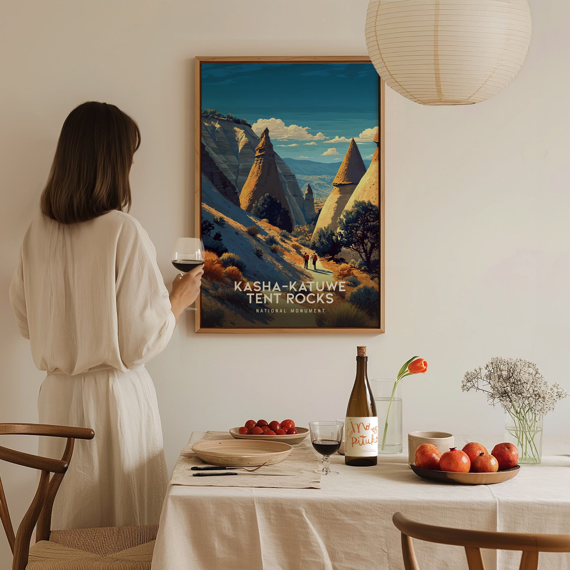 A woman in a white blouse stands at a dining table, holding a glass of wine and admiring a framed art piece of Kasha-Katuwe Tent Rocks National Monument.
