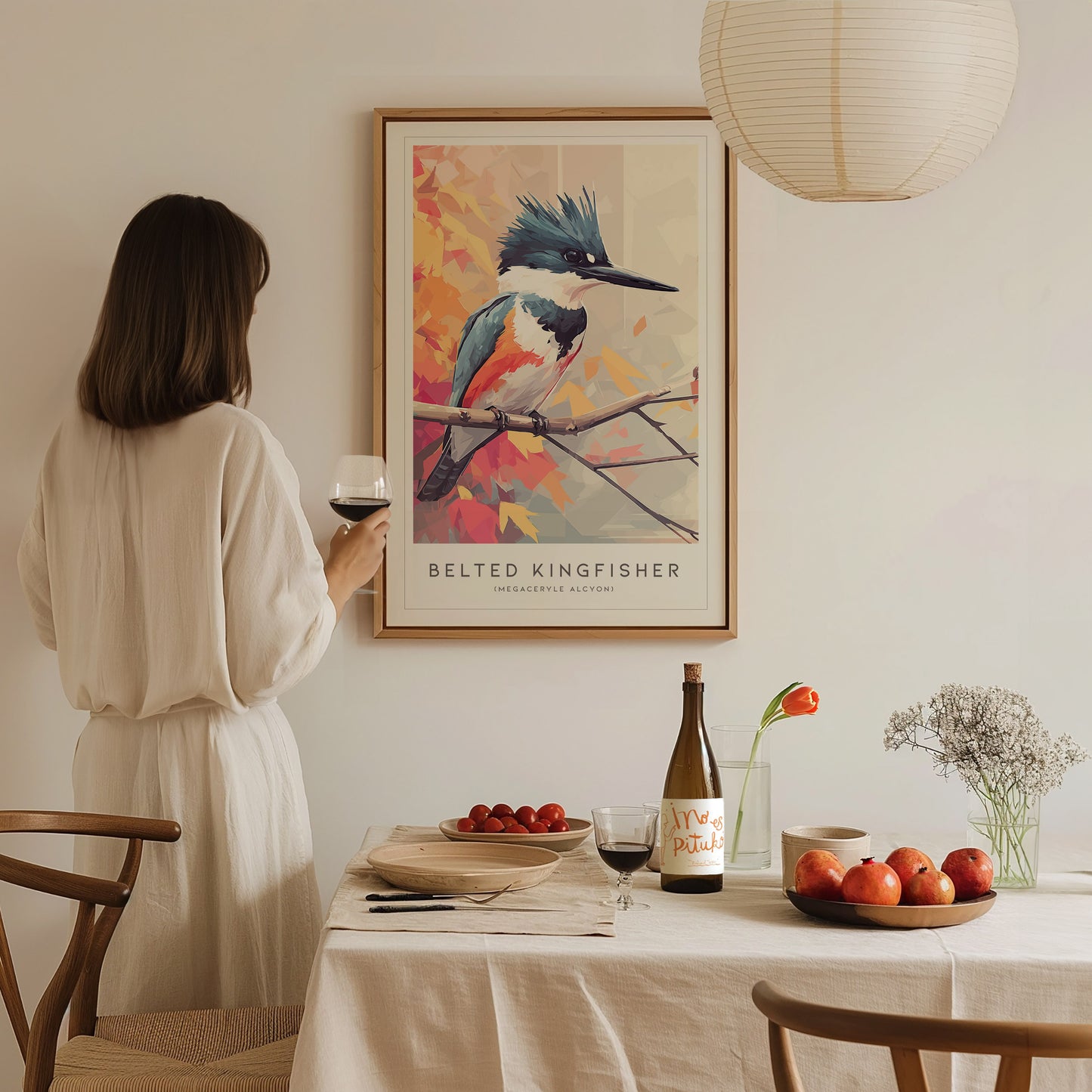 A woman in a light-colored outfit stands at a dining table, holding a glass of wine, while gazing at a colorful art print of a Belted Kingfisher hanging on the wall. The table is set with food and a bottle, creating a cozy atmosphere.