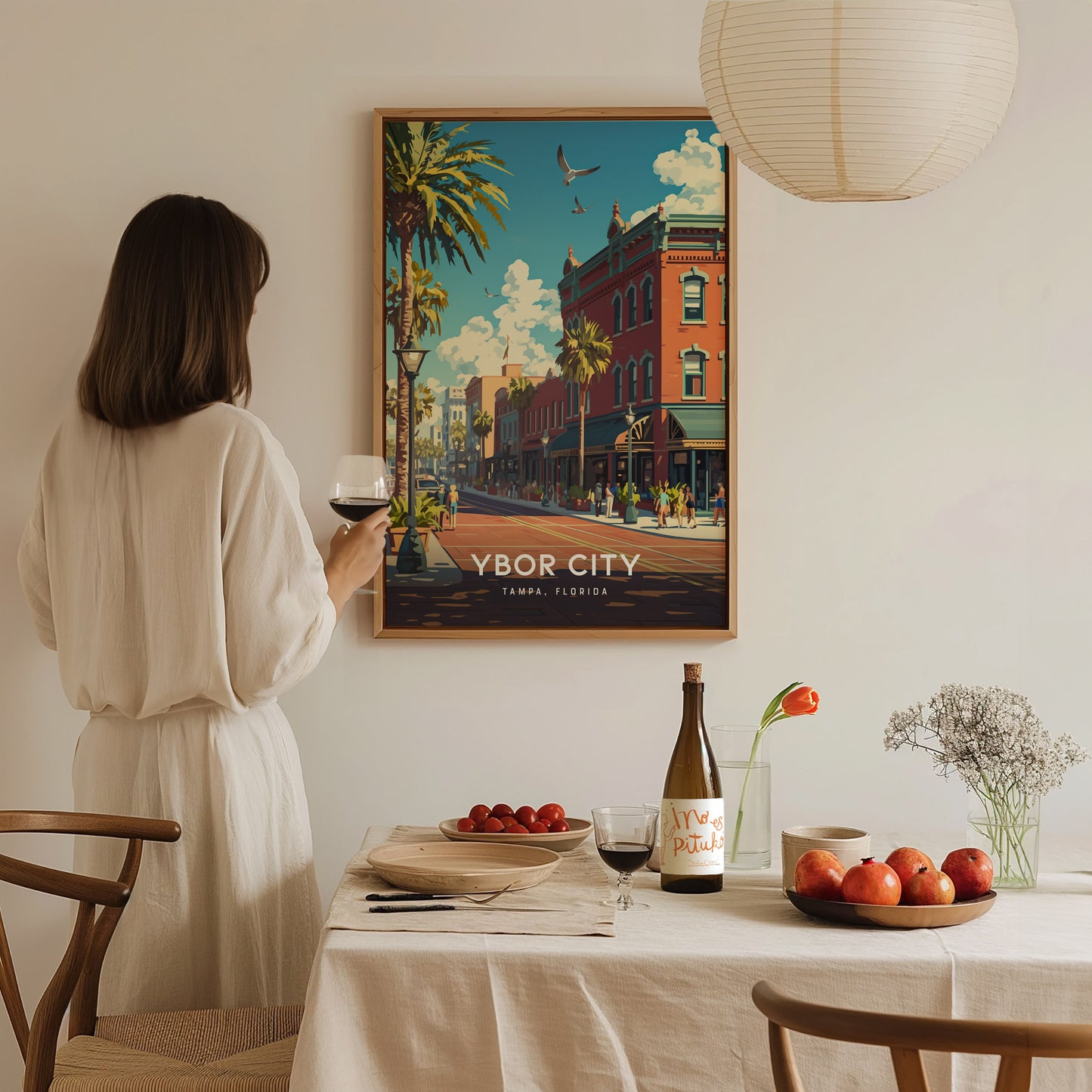 A woman in a white dress admires a colorful Ybor City poster on the wall while holding a glass of wine, with a table set with fruits and flowers.