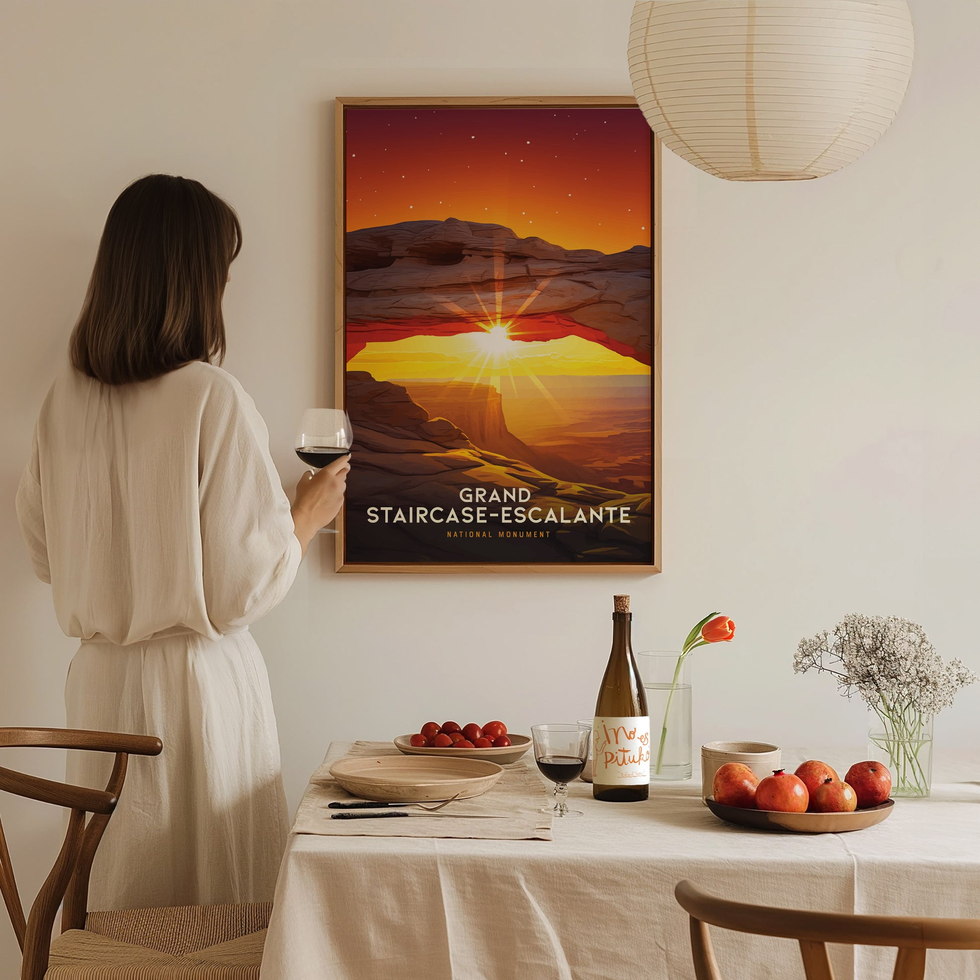 A person in a white dress admires a framed Grand Staircase–Escalante poster. A table with wine, fruit, and flowers is set in the foreground.