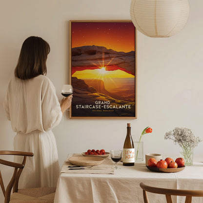 A person in a white dress admires a framed Grand Staircase–Escalante poster. A table with wine, fruit, and flowers is set in the foreground.