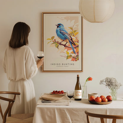 A woman in a light-colored dress stands by a table set with fruit and a bottle, admiring a framed art print of an Indigo Bunting bird perched on a branch. The warm, inviting room features soft lighting and a minimalist decor style.