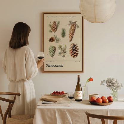 A woman in a white dress holds a glass of wine while admiring a framed pinecone botanical poster. A dining table with fruit, wine, and flowers is in the foreground.